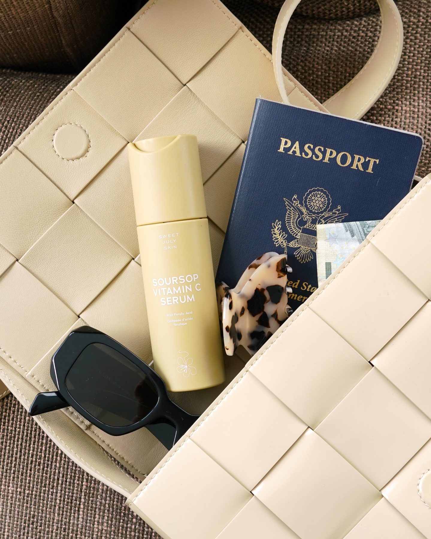 US passport with euro bills, sunglasses, a hair clip and a serum bottle in a cream woven bag
